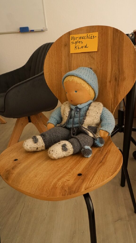 Eine Puppe sitzt auf einem Stuhl. Der Stuhl ist mit einem Zettel, auf dem "Vernachlässigtes Kind" geschrieben ist, beschriftet.