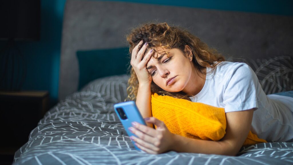 Frau liegt auf Bett und schaut deprimiert aufs Handy. Sie sucht nach Partner*in z.B. bei Tinda